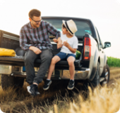 A father and son sit on the tailgate of a pickup truck parked in a field.